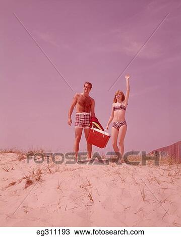 1960S Beach Couple Walking In Sand Carrying Cooler Woman Waving View Large Photo Image Stock Image - 1960S Beach Couple Walking In Sand Carrying Cooler Woman Waving. Fotosearch