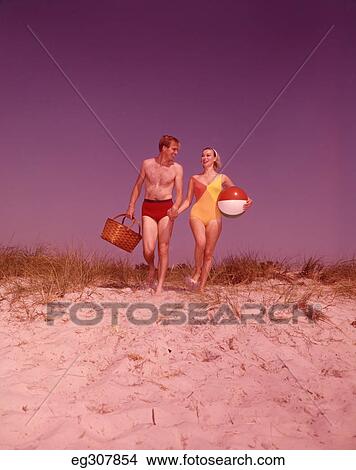 Picture - 1960S Couple Bathing Suits Carrying Picnic Basket On Sand Dune. Fotosearch