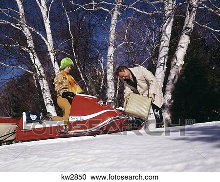 Stock Image - 1960S Couple Man Filling Gas Tank On Red Snowmobile Woman In Orange Outfit & Neon Green Fur Hat At Wheel Snow Birch Tree. Fotosearch