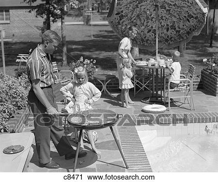 Stock Image - 1960S Family Of 5 On The Patio Father Is Grilling As Daughter Watches Mother Set Umbrella Covered Table . Fotosearch
