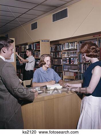 Stock Image - 1960S Four Students In College Library Three At Table And One In Stacks Book Shelf Study Examination Lessons. Fotosearch