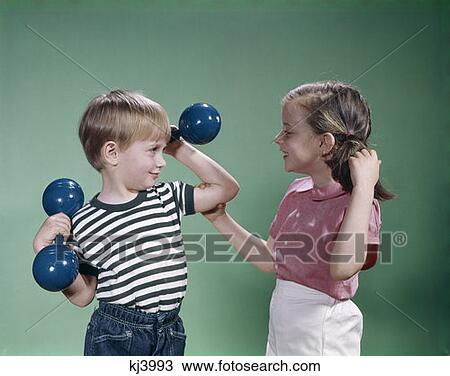 Stock Image - 1960S Little Boy Lifting Hand Weights Little Girl Laughing And Feeling His Muscle Happy Proud Impressed. Fotosearch