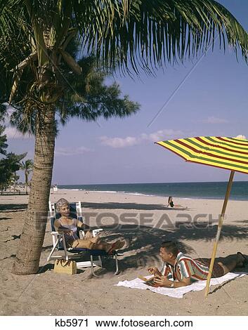 1960S Man And Woman Couple Sunbahing On Beach In Florida Talking Together Under Palm Tree And Colorful Beach Umbrella View Large Photo Image Stock Image - 1960S Man And Woman Couple Sunbahing On Beach In Florida Talking Together Under Palm Tree And Colorful Beach Umbrella. Fotosearch