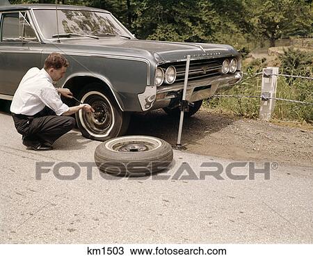 Stock Image - 1960S Man Changing Flat Tire On Car At Side Of Rural Road Car Jack Tools Tire Iron. Fotosearch
