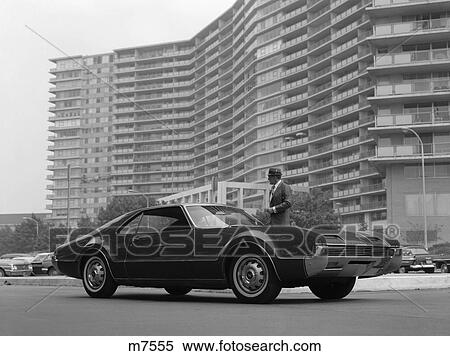 1960S Man In Suit & Hat Standing Beside Oldsmobile Toronado With Large Apartment Building In Background View Large Photo Image Stock Photography - 1960S Man In Suit & Hat Standing Beside Oldsmobile Toronado With Large Apartment Building In Background . Fotosearch