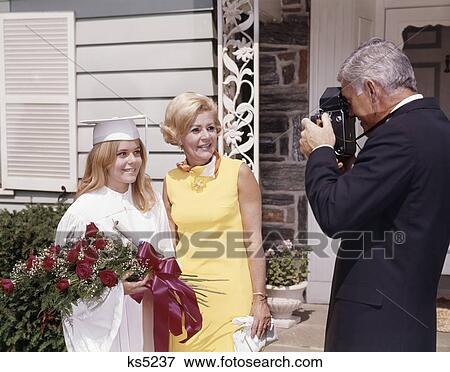 Stock Photo - 1960S Man Taking Picture Of Wife And Daughter In Cap And Gown Outside House Woman Girl Camera Photo Flowers. Fotosearch