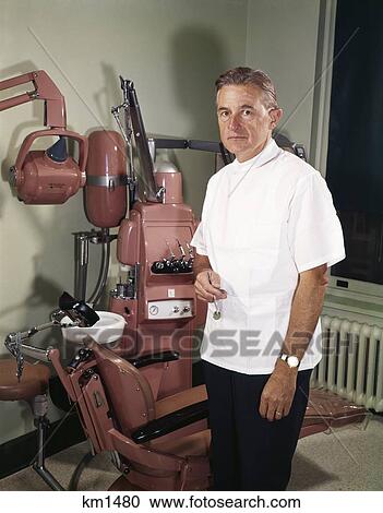 1960S Middle Aged Dentist Wearing White Jacket Standing By Chair In Treatment Room With Dental Drill And Sink View Large Photo Image Stock Image - 1960S Middle Aged Dentist Wearing White Jacket Standing By Chair In Treatment Room With Dental Drill And Sink. Fotosearch