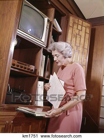 Stock Image - 1960S Middle Aged Woman Selecting Record Albums Long Playing Vinyl Entertainment Center Tv Phonograph Radio Living Room. Fotosearch