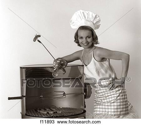 Stock Image - 1960S Portrait Woman Wearing Chef Hat Standing At Bbq Holding A Fork With Hot Dog . Fotosearch