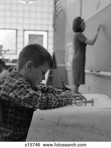 1960S Side View Of Grade School Boy At Desk Writing With Teacher Writing On Blackboard In Soft Focus In Background View Large Photo Image Stock Photograph - 1960S Side View Of Grade School Boy At Desk Writing With Teacher Writing On Blackboard In Soft Focus In Background . Fotosearch