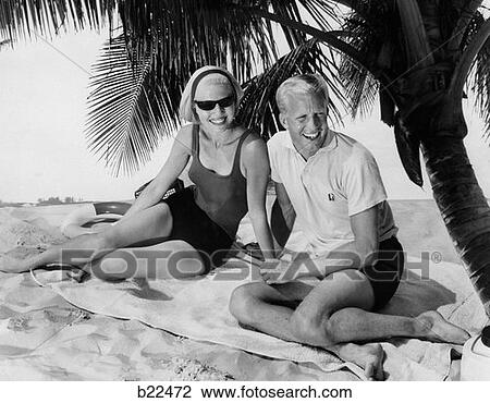 Stock Image - 1960S Smiling Blond Couple Sitting On Beach Towel On Sand Under Palm Tree . Fotosearch