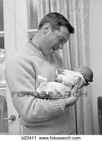 Stock Image - 1960S Smiling Father Holding Newborn Baby In Sleeper . Fotosearch