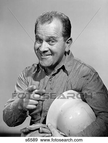Stock Photo - 1960S Smiling Portrait Workman Man Holding Gloves With Hard Hat Under Arm Pointing Index Finger . Fotosearch