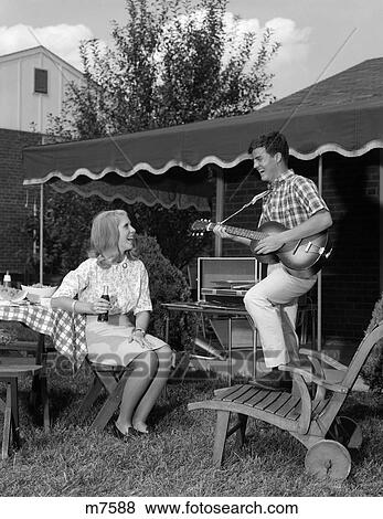 1960S Teenage Couple Man Plays Guitar Woman Sits Holding Soft Drink Bottle Outside Backyard Lawn Furniture View Large Photo Image Stock Photo - 1960S Teenage Couple Man Plays Guitar Woman Sits Holding Soft Drink Bottle Outside Backyard Lawn Furniture . Fotosearch