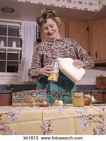 Stock Image - 1960S Woman Mom Kitchen Pack Make School Lunch Metal Lunchbox Thermos Pour Milk Green Apron Print Shirt Smile Housewife. Fotosearch