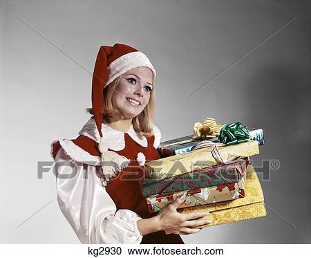Stock Image - 1960S Young Woman In Red And White Santa Helper Costume And Hat Holding Pile Of Wrapped Christmas Presents Studio. Fotosearch
