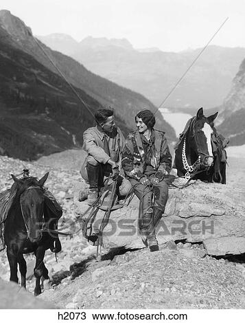 Stock Image - 1920s 1930s couple man woman wearing riding gear jodhpurs boots spurs sitting on large rock by two horses with western saddles. Fotosearch