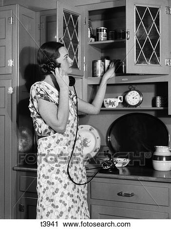 1920s 1930s woman housewife wearing apron in kitchen talking on telephone putting canister on cupboard shelf View Large Photo Image Stock Image - 1920s 1930s woman housewife wearing apron in kitchen talking on telephone putting canister on cupboard shelf. Fotosearch