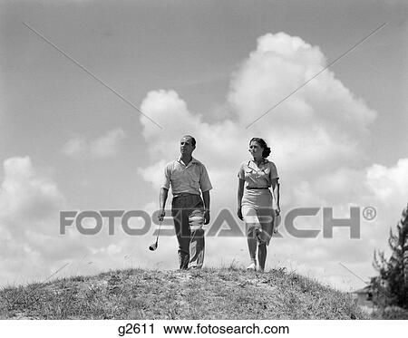 Stock Image - 1930s couple man woman golfers walking together over crest of hill. Fotosearch