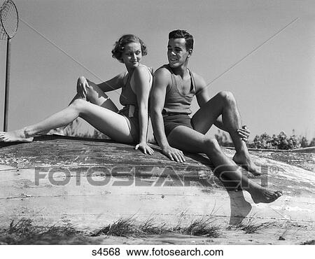 1930s couple man woman smiling wearing bathing suits sitting back to back on overturned boat hull View Large Photo Image Stock Photo - 1930s couple man woman smiling wearing bathing suits sitting back to back on overturned boat hull. Fotosearch
