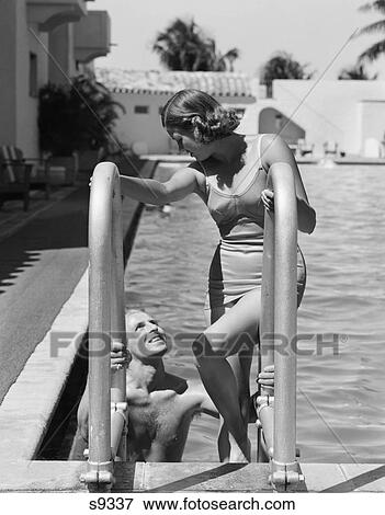 Stock Photo - 1930s couple woman wearing one piece bathing suit climbing ladder out of swimming pool looking down at man smiling miami beach florida. Fotosearch