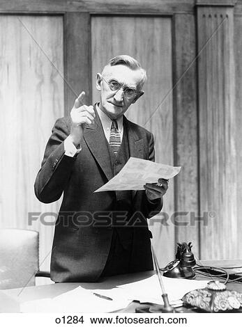 Picture - 1930s elderly man in office standing behind desk gesturing with raised finger. Fotosearch