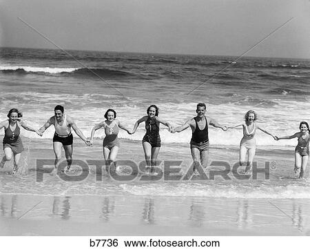 Stock Photograph - 1930s group 7 people holding hands running out of surf onto beach. Fotosearch
