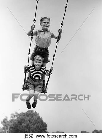 Stock Image - 1930s two smiling boys sitting standing on playground swing. Fotosearch
