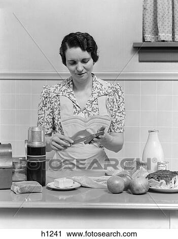 Stock Image - 1930s woman housewife in kitchen wearing apron making sandwich packing metal lunch box thermos apples bread milk on counter. Fotosearch