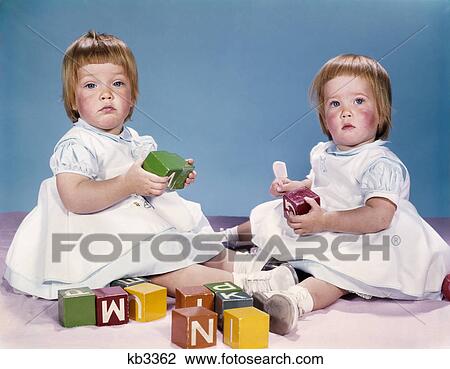 Stock Image - 1950s 1960s portrait two twin baby girls wearing dresses playing with toy blocks. Fotosearch