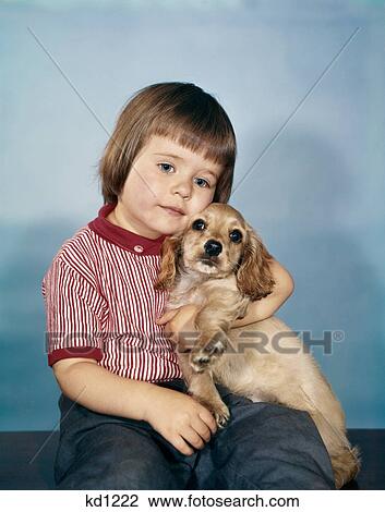 Stock Image - 1950s 1960s winsome sad little girl sitting hugging a cocker spaniel puppy studio. Fotosearch