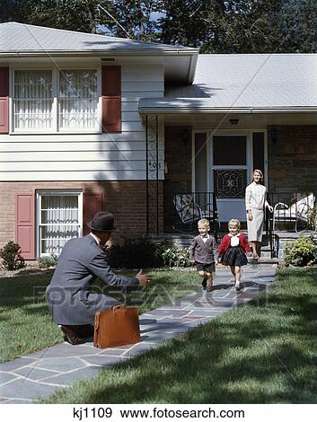 Stock Photo - 1950s family woman mother watching man father coming home to suburban house kneeling arms extended to boy son girl daughter running to welcome him home. Fotosearch