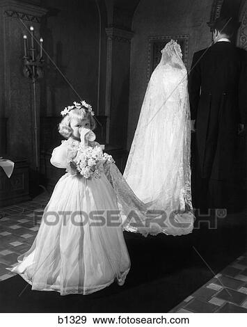 Stock Photo - 1950s little girl bridesmaid drinking glass of milk holding bridal veil train walking down church aisle looking at camera. Fotosearch