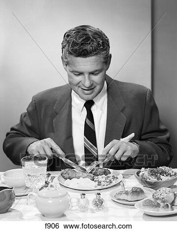 1950s smiling man in suit and tie sitting at table holding knife and fork eating dinner or supper meal View Large Photo Image Stock Photograph - 1950s smiling man in suit and tie sitting at table holding knife and fork eating dinner or supper meal. Fotosearch