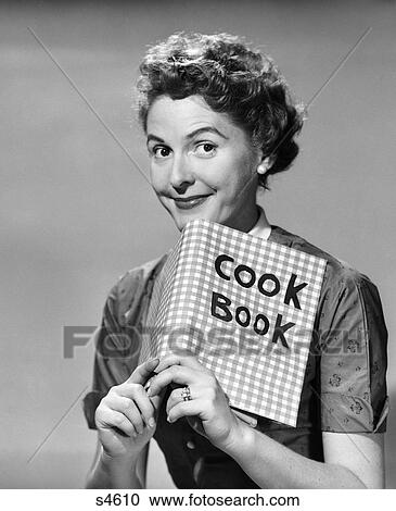 Stock Image - 1950s smiling woman looking over the top of a cook book. Fotosearch