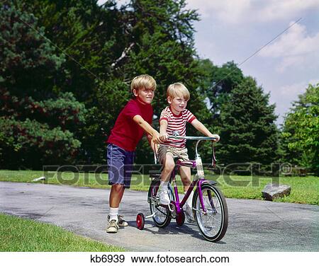1960s 1970s boy helping little brother ride two wheel bicycle with training wheels View Large Photo Image Stock Photo - 1960s 1970s boy helping little brother ride two wheel bicycle with training wheels. Fotosearch