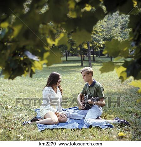 Stock Image - 1960s 1970s smiling couple sitting on blanket on grass man playing acoustic guitar. Fotosearch