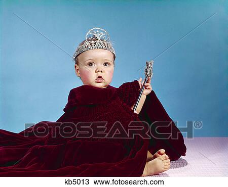 Stock Image - 1960s baby dressed as royal queen velvet robe cloak cape rhinestone tiara crown and scepter wand. Fotosearch