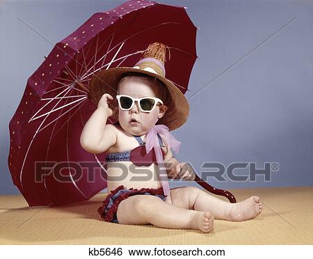 Stock Photograph - 1960s baby girl wearing two piece bikini and straw hat sunglasses sitting by red beach umbrella. Fotosearch