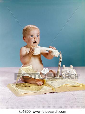Stock Image - 1960s baby surprised facial expression holding telephone hand set over yellow pages phone book directory. Fotosearch