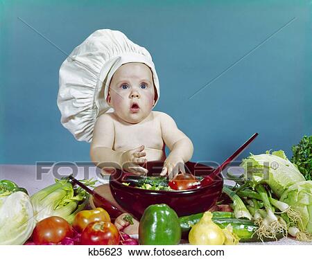 Stock Image - 1960s baby wearing chef hat about to toss salad with hands wooden salad bowl fresh vegetables ingredients. Fotosearch