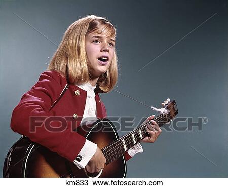 Stock Image - 1960s blond teenaged girl singing and playing acoustic guitar. Fotosearch