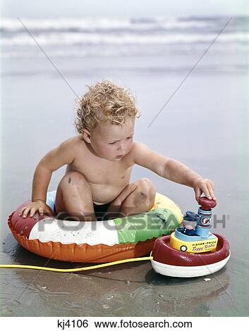 1960s blond toddler sitting in inner tube on beach playing with toy boat View Large Photo Image Stock Photograph - 1960s blond toddler sitting in inner tube on beach playing with toy boat. Fotosearch