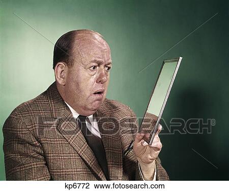 Stock Image - 1960s businessman looking into hand held mirror with serious facial expression. Fotosearch