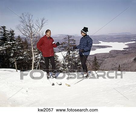 Stock Photo - 1960s couple man woman skiers top hill with winter lake vista view. Fotosearch