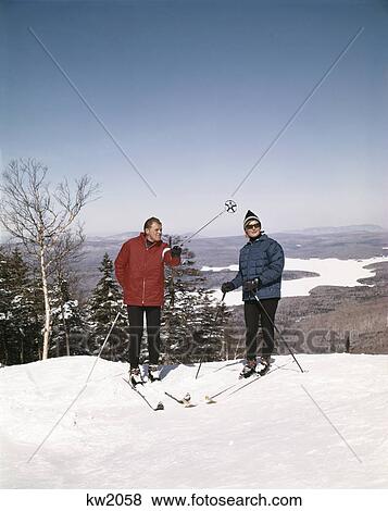 Stock Photo - 1960s couple man woman skiers top hill winter lake vista view. Fotosearch