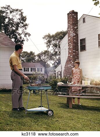 1960s couple suburban backyard man grilling woman setting picnic table View Large Photo Image Stock Photo - 1960s couple suburban backyard man grilling woman setting picnic table. Fotosearch