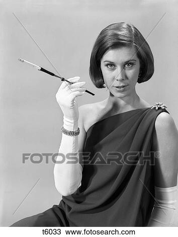 Stock Image - 1960s formally elegantly dressed young woman sexy expression holding long cigarette holder wearing long white gloves. Fotosearch