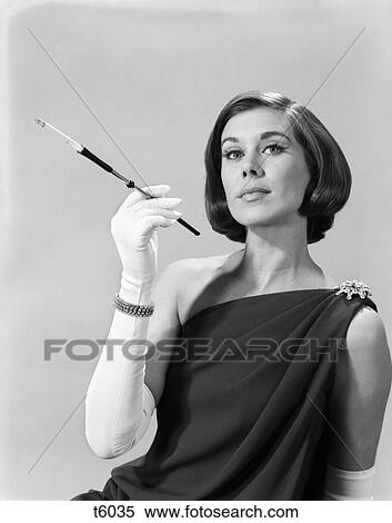 Stock Photography - 1960s formally elegantly dressed young woman arrogant expression holding long cigarette holder wearing long white gloves. Fotosearch
