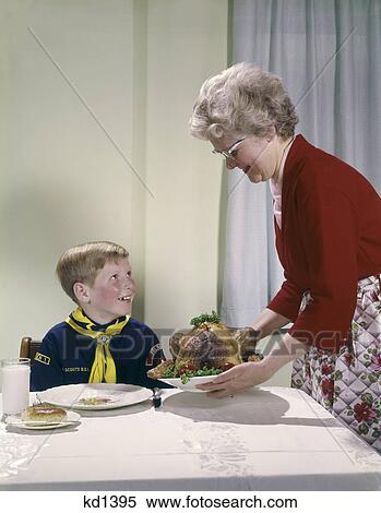 Stock Photography - 1960s grandmother putting turkey on table near boy in cub scout uniform woman grandson feast happy occasion food. Fotosearch